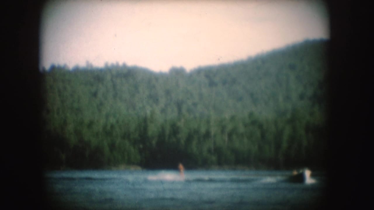 Water skier rides across a forest-lined South Dakota lake as a tow boat pulls ahead, filmed on 8mm in 1954.