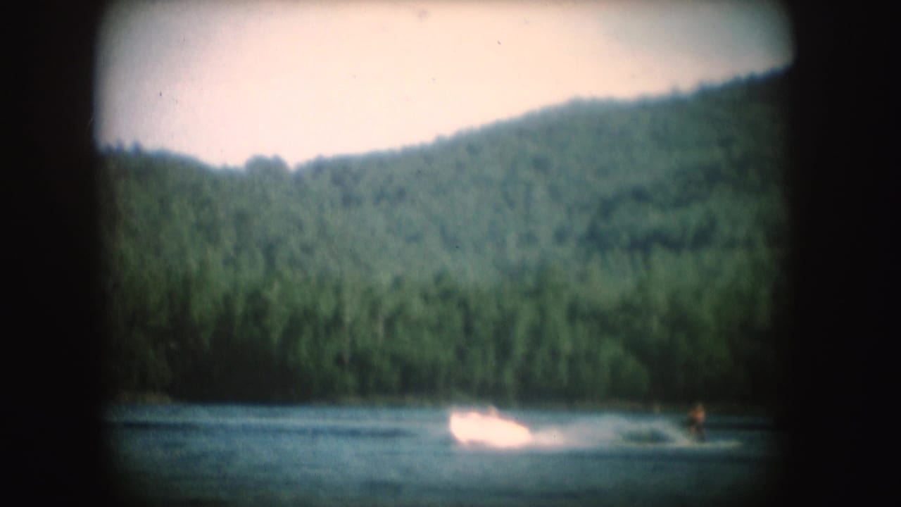 1954 South Dakota lake scene with a small motorboat throwing a bright wake as a distant skier moves behind it.