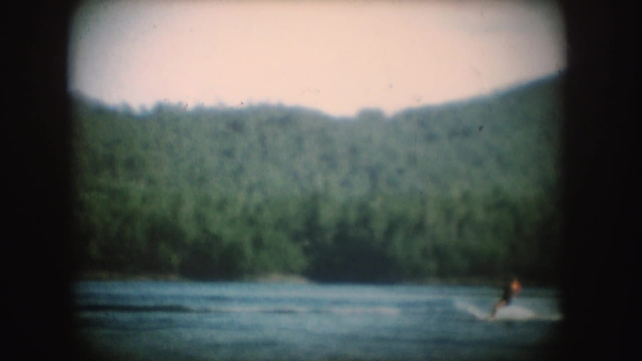 1954 South Dakota home-movie frame with a distant person water skiing across a lake, leaving a white wake near the wooded shoreline.