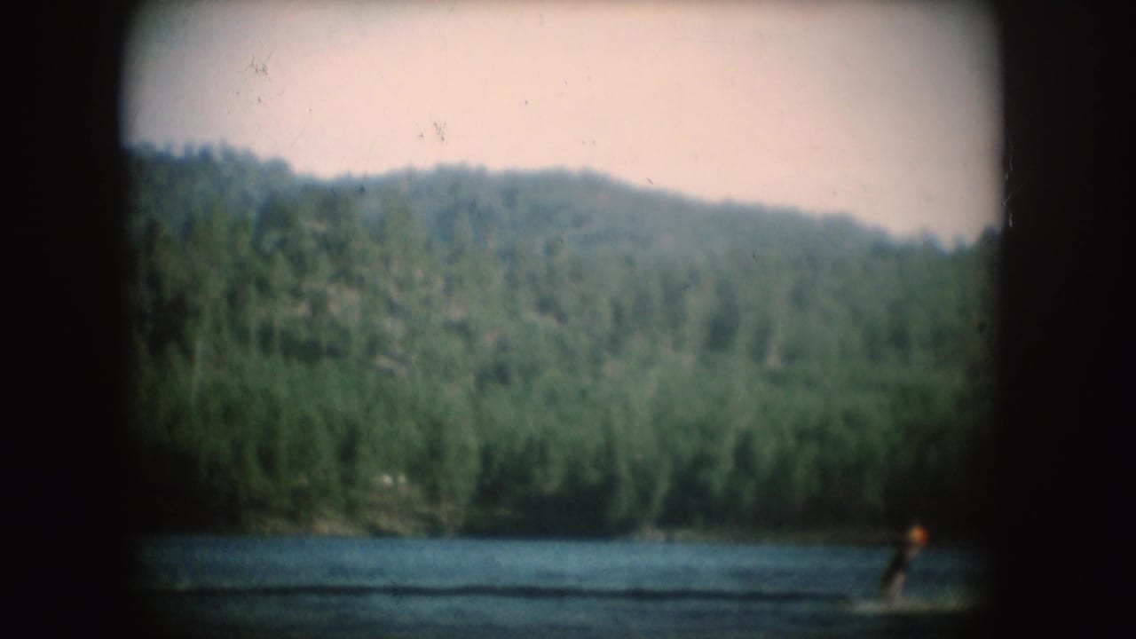 1954 South Dakota 8mm frame showing a small tow boat and distant water skier on a lake bordered by dense trees and hills.