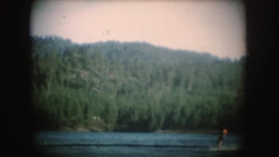 1954 South Dakota 8mm frame showing a small tow boat and distant water skier on a lake bordered by dense trees and hills.