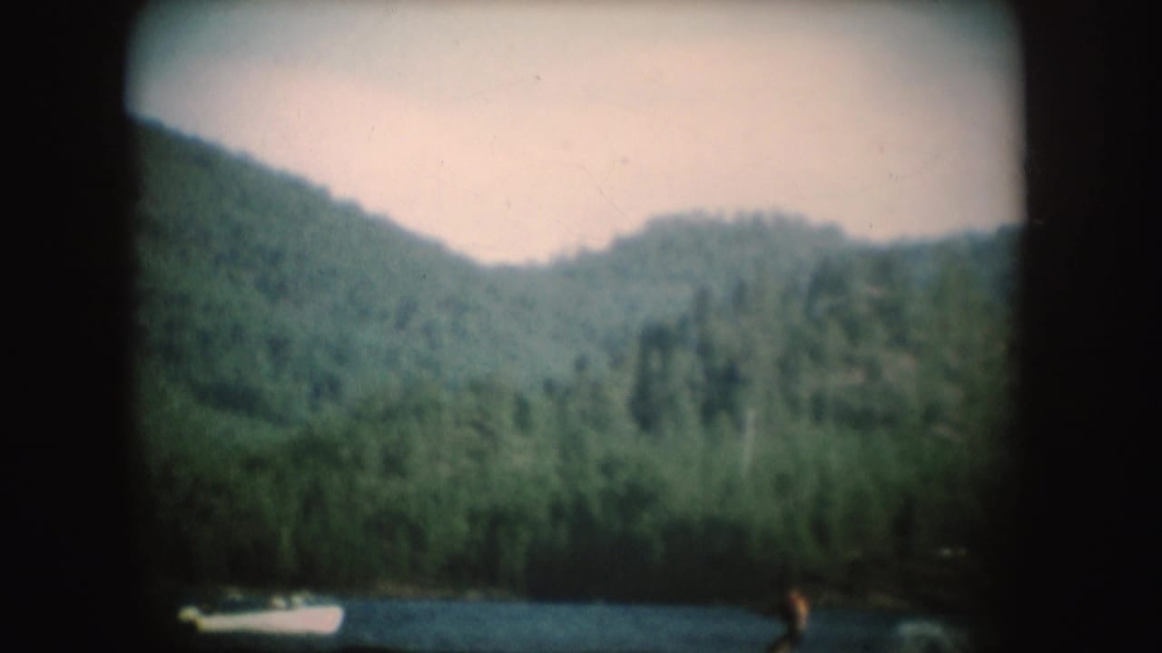 1954 South Dakota home-movie frame with a small white boat towing a distant water skier across a wooded lake setting.