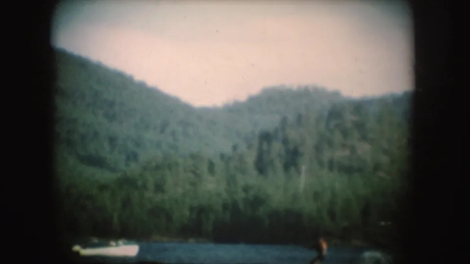 1954 South Dakota home-movie frame with a small white boat towing a distant water skier across a wooded lake setting.