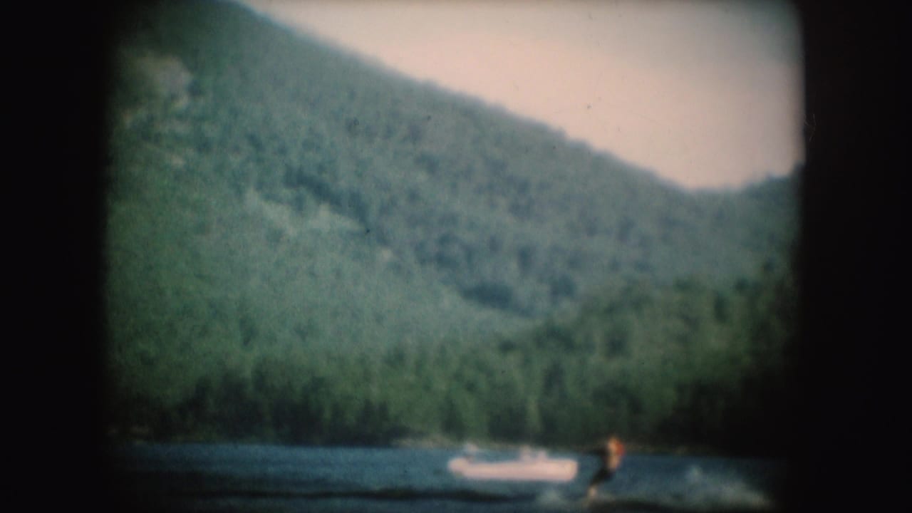 1954 South Dakota 8mm frame showing a distant water skier crossing a lake below wooded hills, with a small boat nearby.