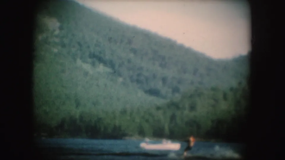 1954 South Dakota 8mm frame showing a distant water skier crossing a lake below wooded hills, with a small boat nearby.