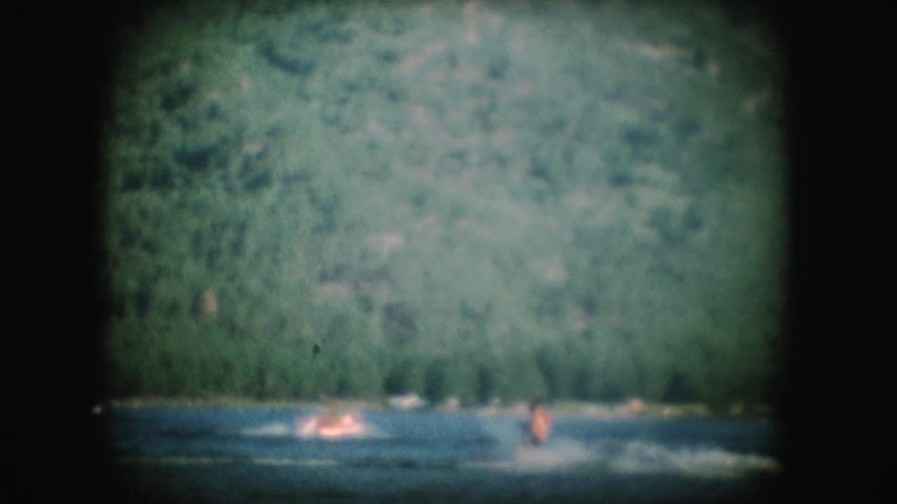 A 1954 South Dakota home-movie frame shows a water skier kicking up spray across a lake below green hills.