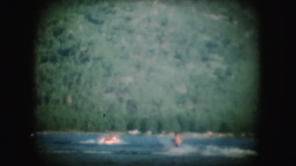 A 1954 South Dakota home-movie frame shows a water skier kicking up spray across a lake below green hills.
