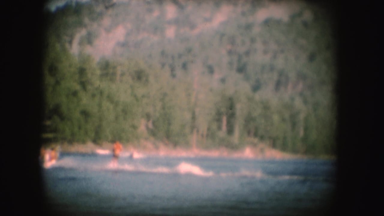 1954 South Dakota 8mm frame with a distant water skier in an orange vest leaving a white wake near a wooded lakeshore.