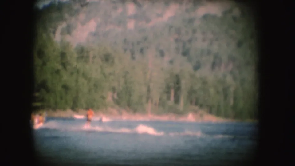 1954 South Dakota 8mm frame with a distant water skier in an orange vest leaving a white wake near a wooded lakeshore.