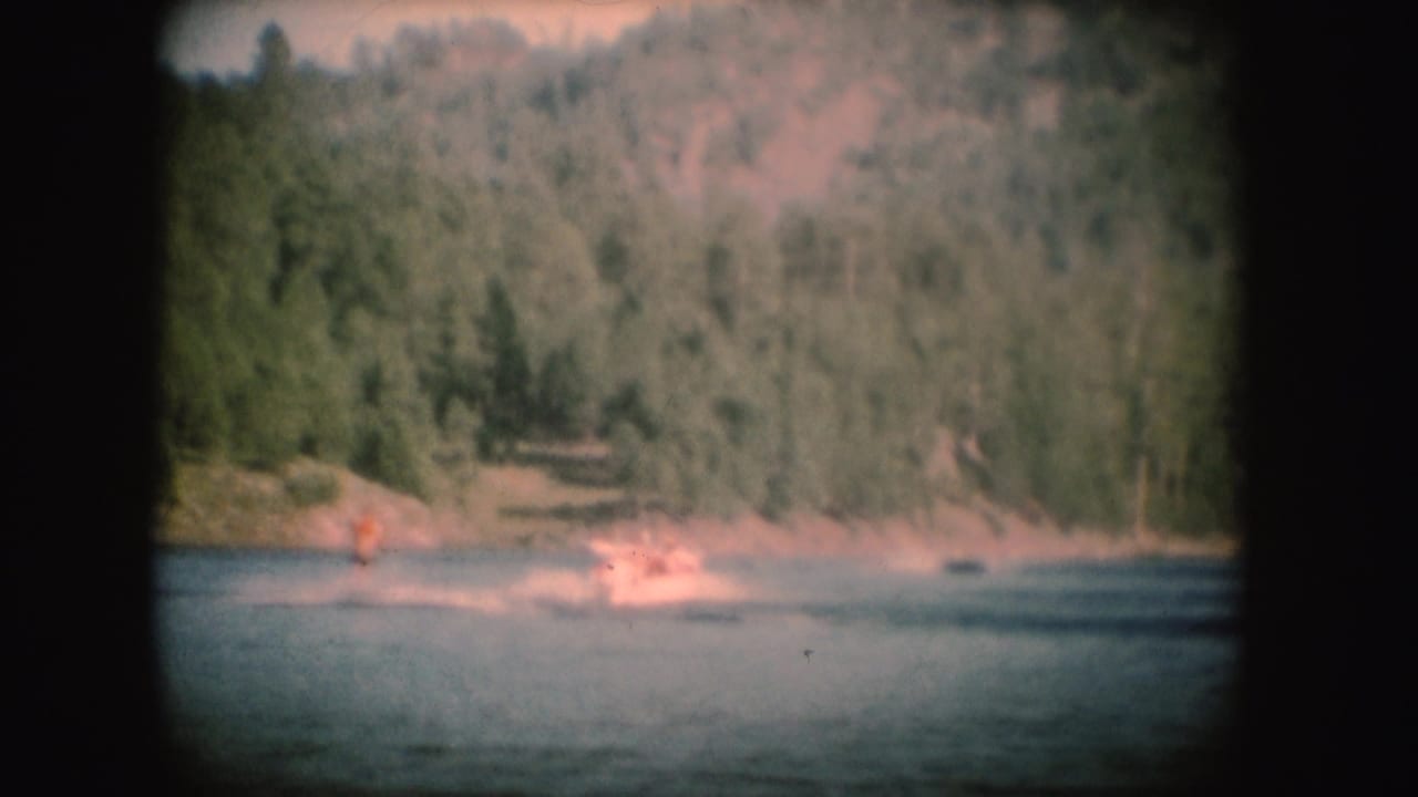 Distant 1954 South Dakota lake view showing a water skier and bright wake against a wooded shoreline.