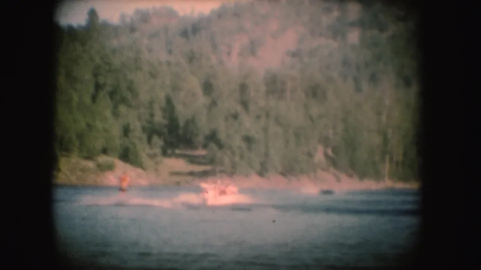 Distant 1954 South Dakota lake view showing a water skier and bright wake against a wooded shoreline.