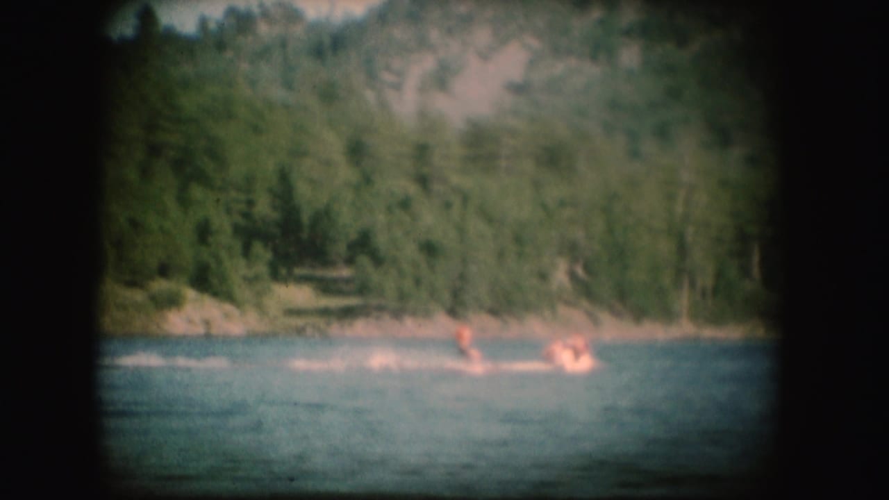 1954 South Dakota home-movie frame of a water skier in orange crossing the lake before a wooded hillside.