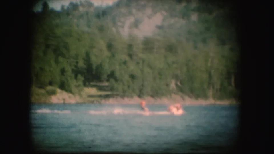 1954 South Dakota home-movie frame of a water skier in orange crossing the lake before a wooded hillside.
