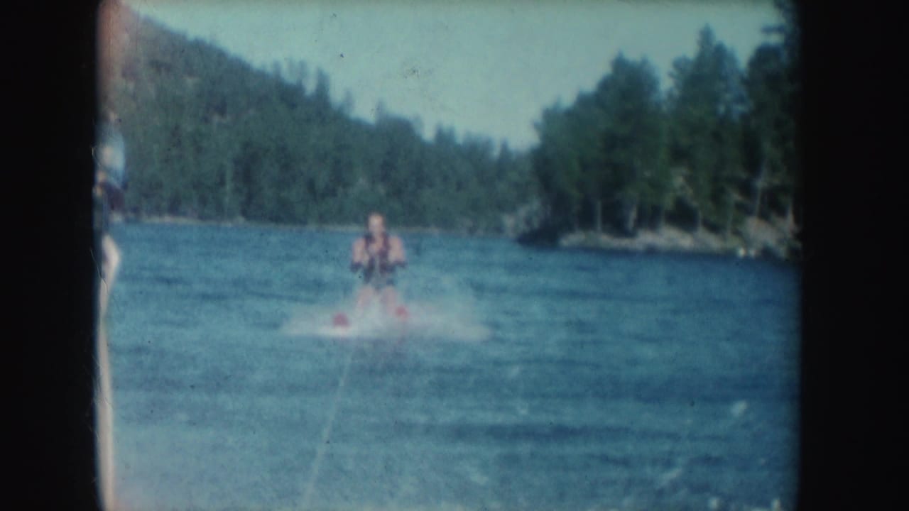 A water skier is towed across a South Dakota lake in 1954 home-movie footage, with spray on the water and trees along the shore.