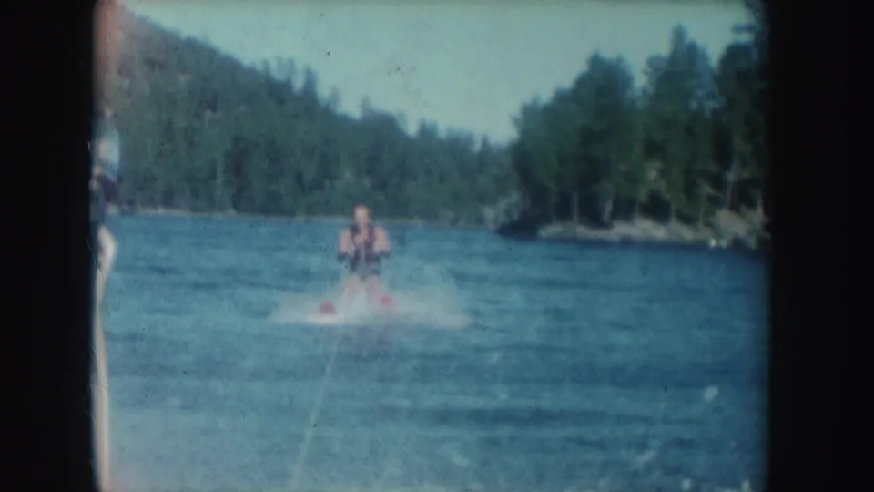 A water skier is towed across a South Dakota lake in 1954 home-movie footage, with spray on the water and trees along the shore.