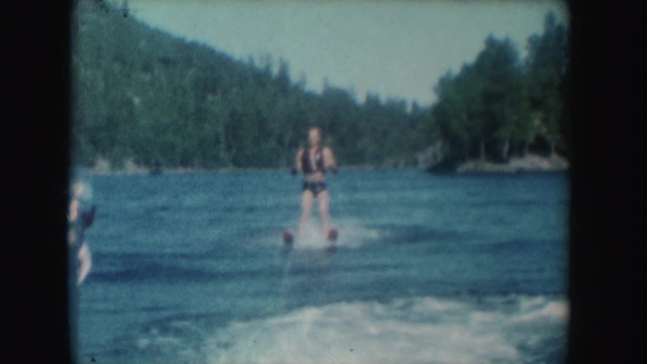 Water skier follows a boat across a lake with forested hills behind him.
