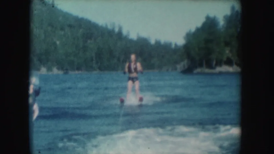 1954 South Dakota home-movie frame showing a man water skiing behind a boat on a blue lake with wooded hills in the background.
