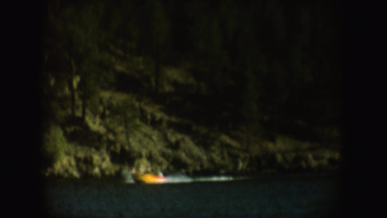Small lit boat moving across dark water below a forested shoreline.