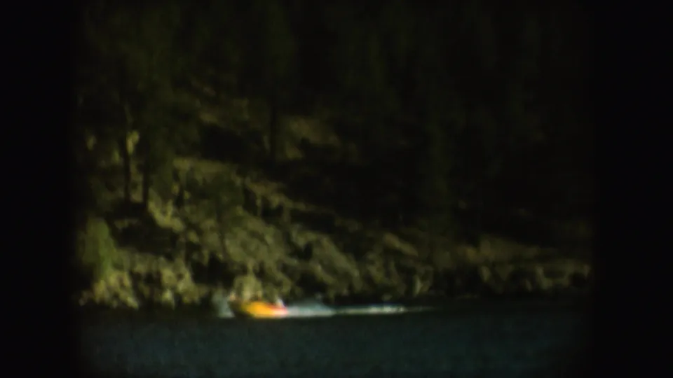 1954 South Dakota 8mm frame with a small lighted boat on dark river water beneath trees and rocky bank.