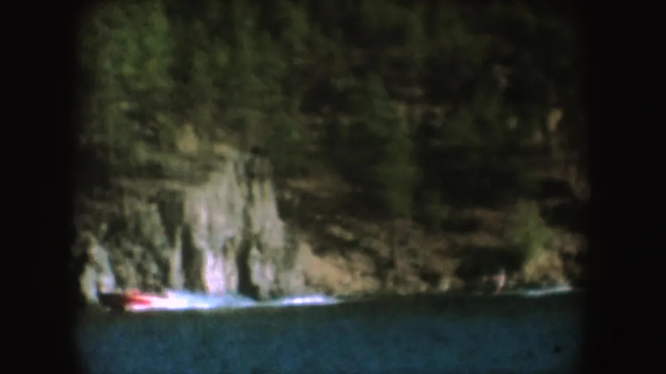 South Dakota lake recreation frame with a red motorboat and white wake near a wooded rocky shore, from 1954 8mm home-movie footage.