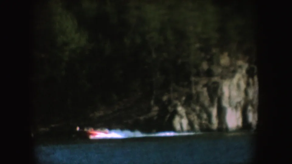A small red boat crosses dark lake water near a wooded rock face, leaving a bright wake in a 1954 South Dakota 8mm frame.