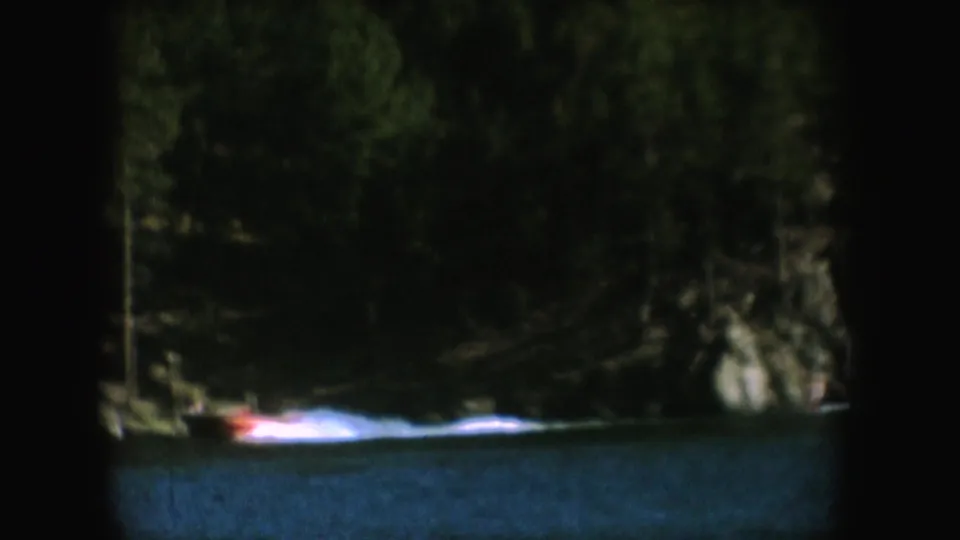 A small speedboat crosses a wooded South Dakota lake in 1954 home-movie footage, with soft focus and dark film edges.