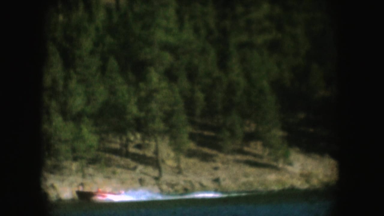 The boat passes low in frame against a forested bank, with the 1954 South Dakota lake scene softened by 8mm grain.