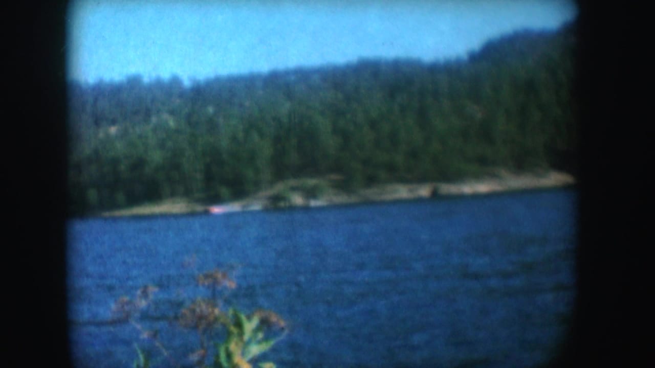 A wider South Dakota lake view shows blue water, wooded hills, sky, and a small distant boat in 1954 home-movie footage.