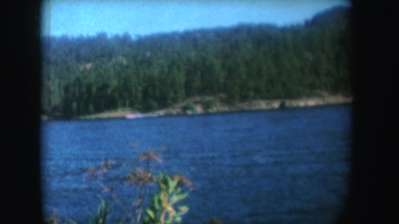Blue lake with forested shoreline, low hills, and a distant red boat seen through dark 8mm film borders.