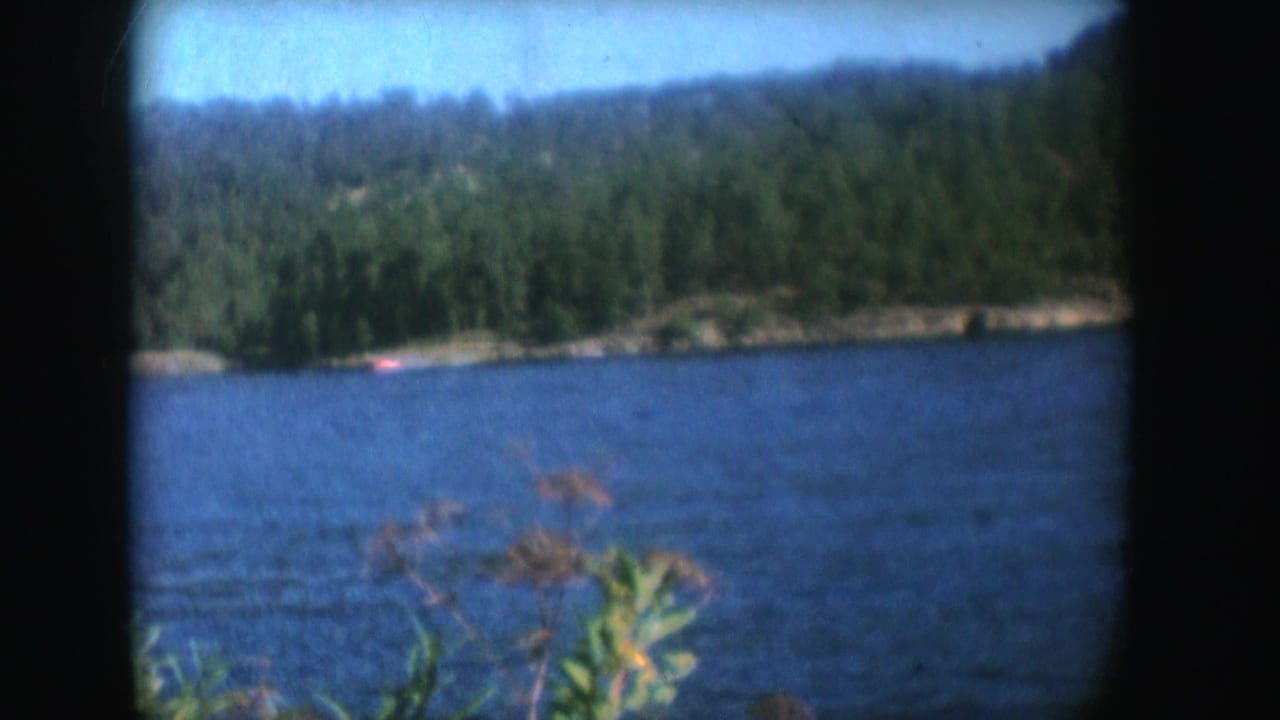 1954 South Dakota home-movie frame of a distant red motorboat on a blue lake with trees and hills beyond.