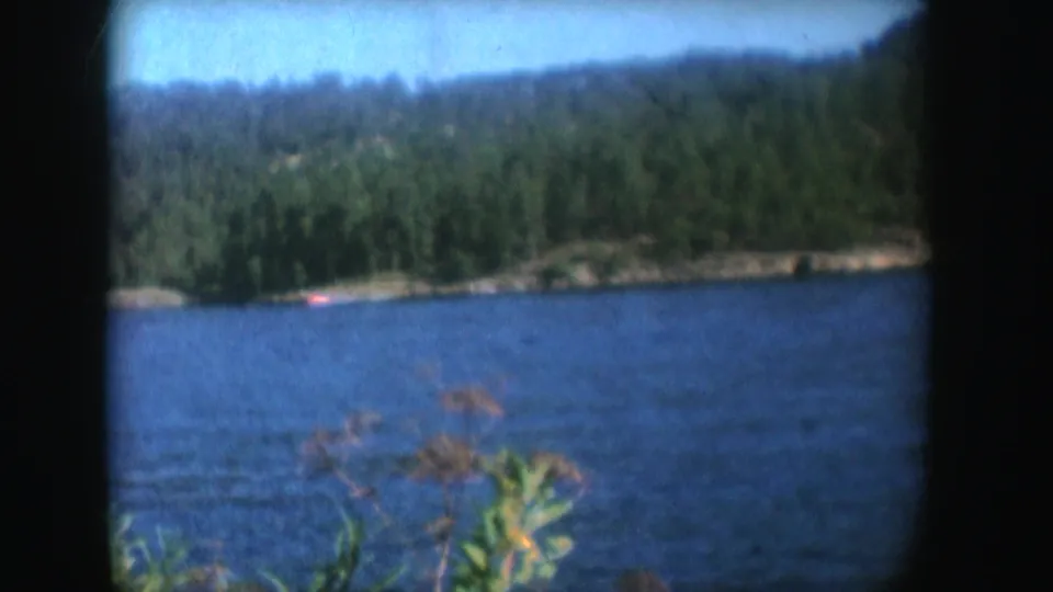 1954 South Dakota home-movie frame of a distant red motorboat on a blue lake with trees and hills beyond.