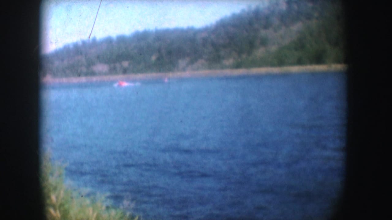1954 South Dakota 8mm still with a red boat near a distant shoreline, blue water, hazy hills, and soft film color.