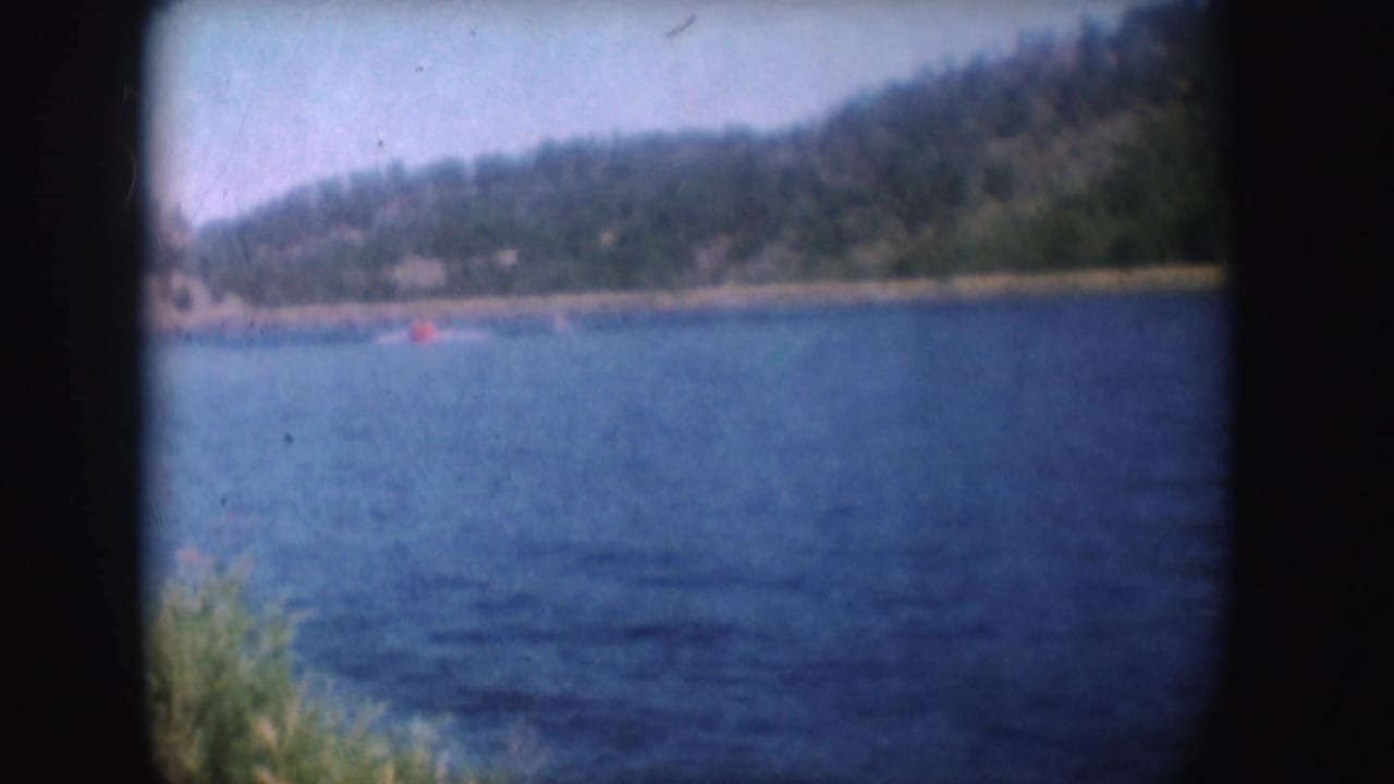 1954 South Dakota home-movie frame showing the red boat across the lake with wooded hills and soft 8mm exposure.