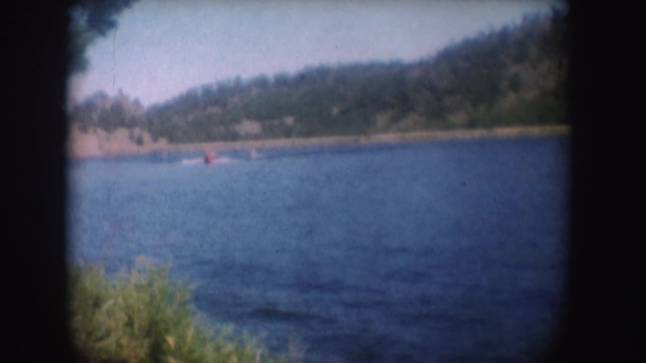 1954 South Dakota home-movie frame of a small red speedboat crossing a blue lake beneath a wooded shoreline.