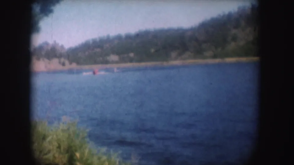 1954 South Dakota home-movie frame of a small red speedboat crossing a blue lake beneath a wooded shoreline.