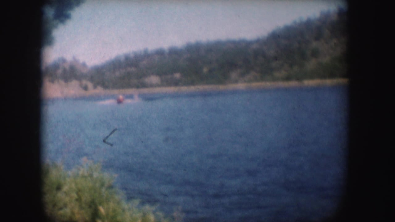 1954 South Dakota 8mm still with a red speedboat low on blue lake water, distant trees, and soft home-movie focus.