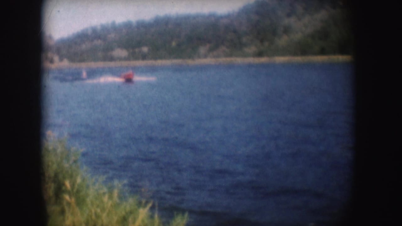 1954 South Dakota frame showing the red speedboat leaving a white wake across blue lake water near a wooded shoreline.