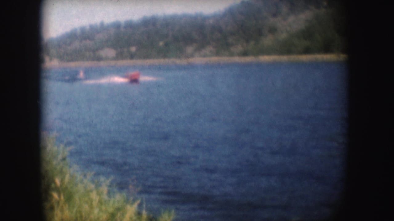Red speedboat far across blue water with trees and hillside behind it.