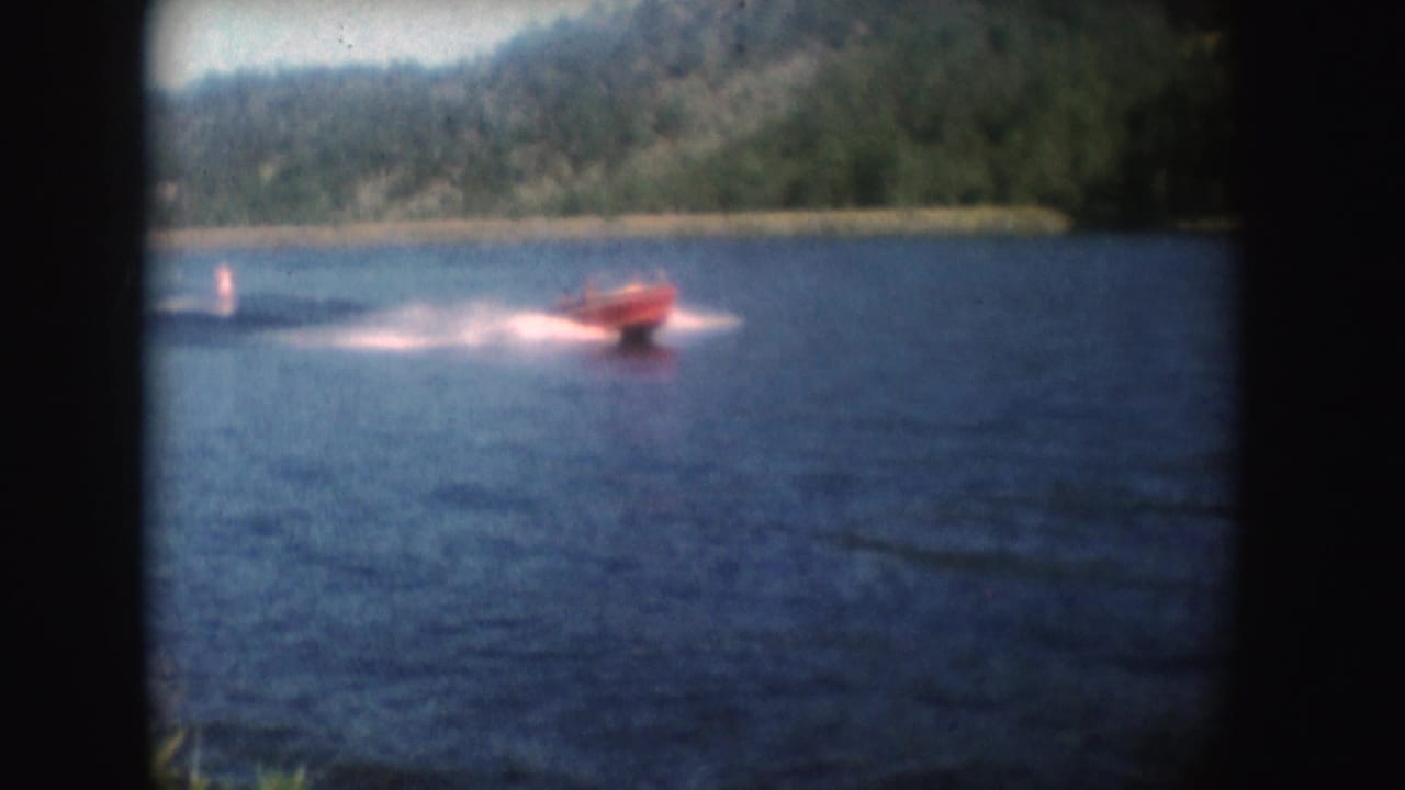 A red speedboat passes closer to the shore in 1954 South Dakota footage, throwing a white wake across blue water.