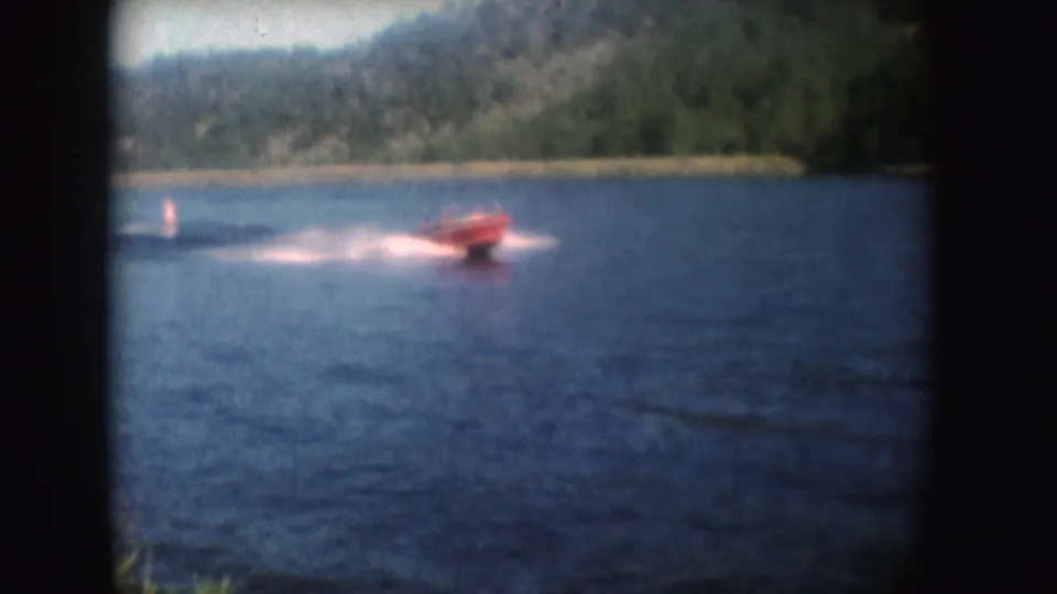 A red speedboat passes closer to the shore in 1954 South Dakota footage, throwing a white wake across blue water.