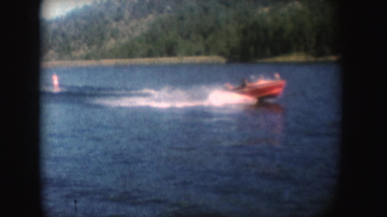 Close shoreline view of a red speedboat on South Dakota water in 1954, with a broad foamy wake and trees behind.