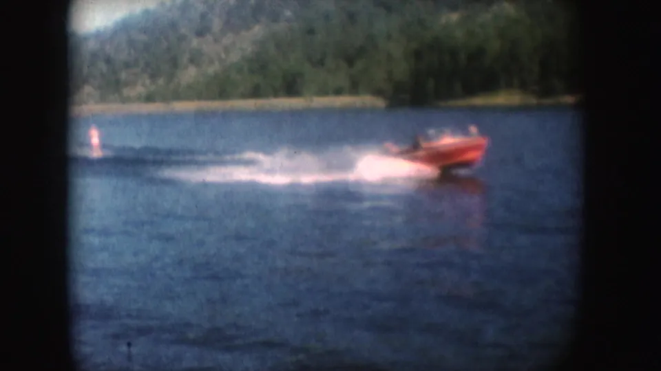 Close shoreline view of a red speedboat on South Dakota water in 1954, with a broad foamy wake and trees behind.