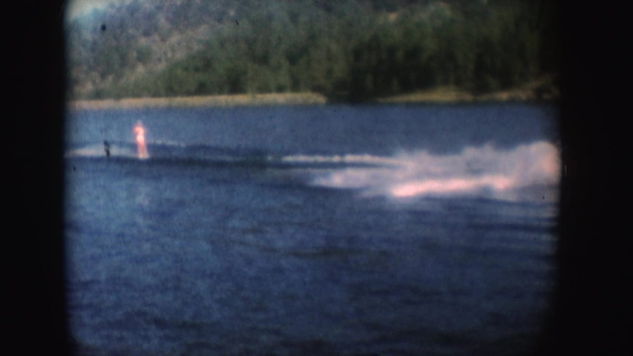 1954 South Dakota 8mm frame showing a distant water skier crossing a sunlit lake with a large spray wake and wooded shoreline.