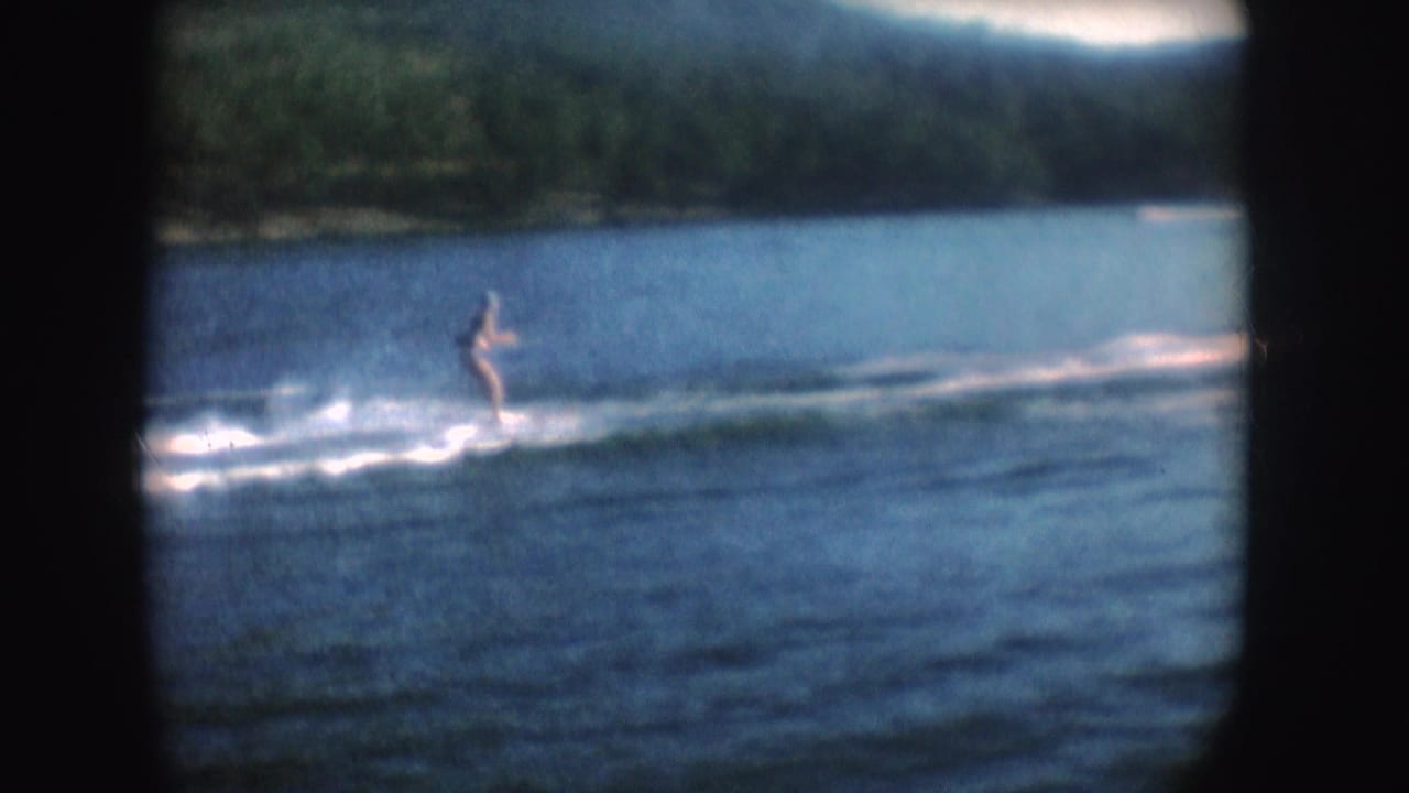 1954 South Dakota 8mm frame showing a distant skier bending low on a sunlit lake, with wake foam and wooded shoreline in view.