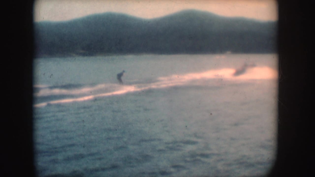 1954 South Dakota 8mm frame showing a small distant skier on a broad lake, with wake spray, hazy hills, and dark frame borders.