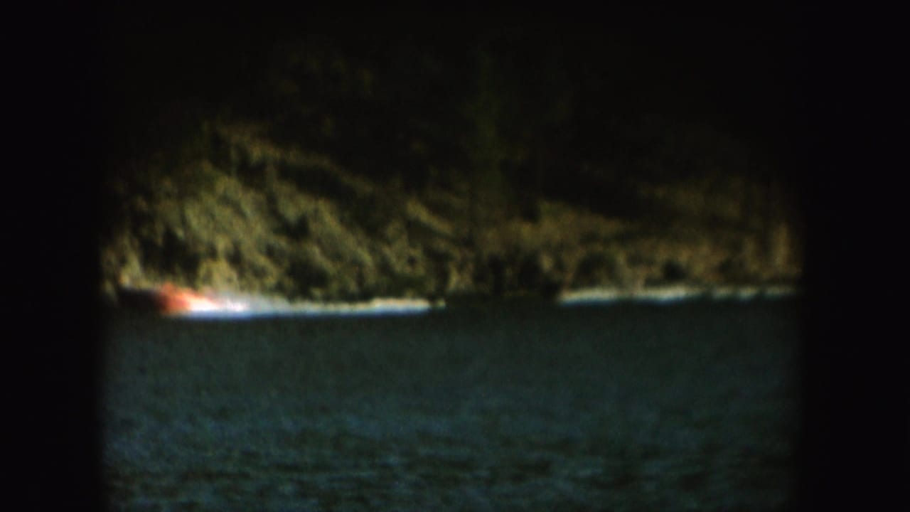 A 1954 South Dakota lake scene shows a boat towing a skier past trees and rocky shoreline on grainy 8mm film.