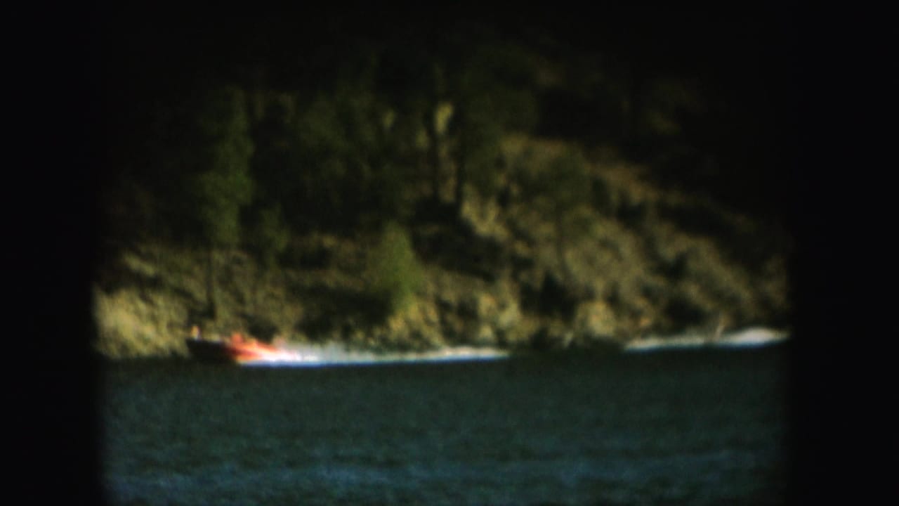 Grainy 1954 South Dakota home-movie frame of a motorboat pulling a water skier across a lake beneath wooded hills.