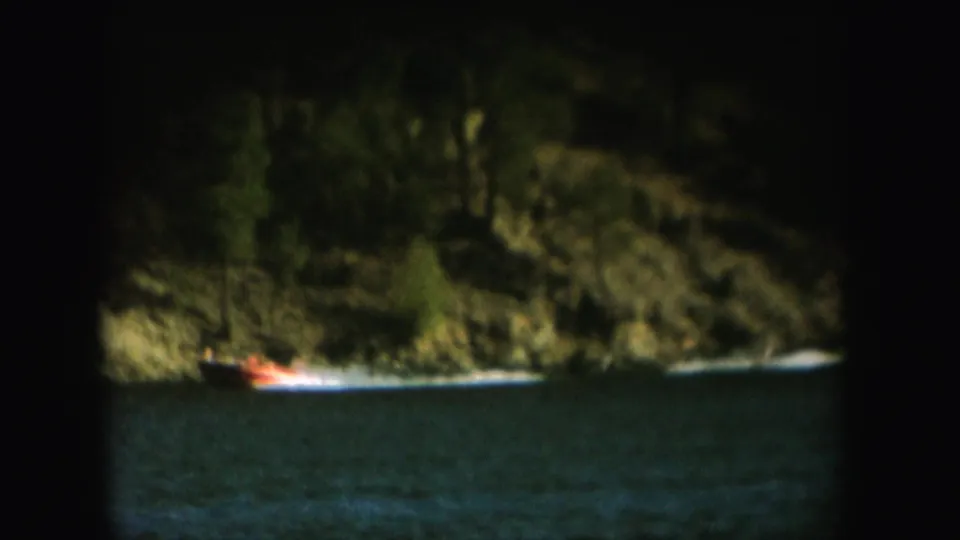 Grainy 1954 South Dakota home-movie frame of a motorboat pulling a water skier across a lake beneath wooded hills.