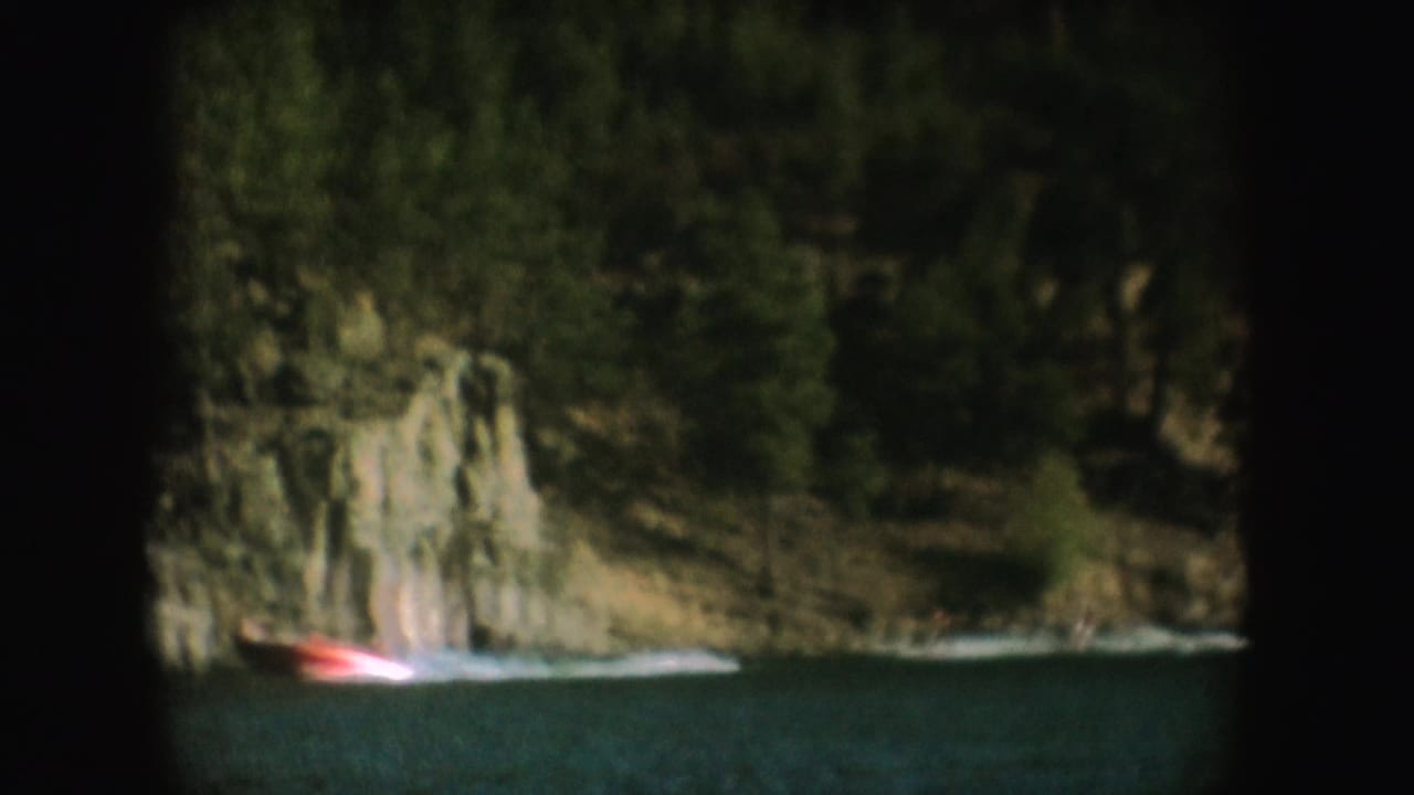 A distant boat and water skier pass a rocky, wooded South Dakota shoreline in a dark-edged 1954 8mm frame.