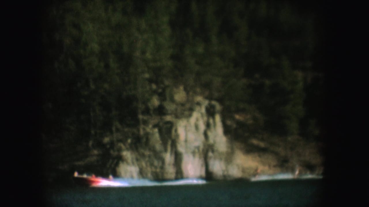 1954 South Dakota 8mm frame showing a small boat crossing shaded lake water near rocks and dense trees.